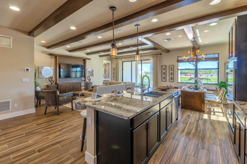 A modern open-concept kitchen and living area at Villamar at Toscana Isles features dark wooden cabinets, a granite island with stools, and wood-patterned flooring. The living area boasts a large TV and seating, with a view of the patio through sliding glass doors.