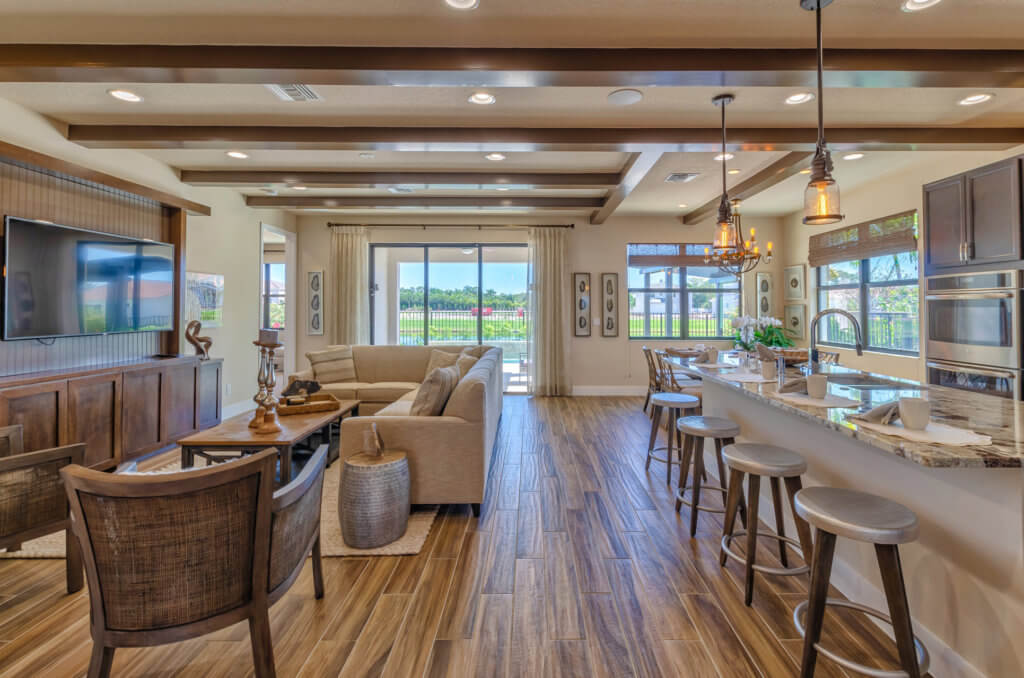 A modern open-concept living area at Villamar at Toscana Isles combines a kitchen with bar stools, a dining space, and a living room featuring a sectional couch. Large windows and glass doors offer natural light and outdoor views. Neutral tones and wood accents enhance the elegance.