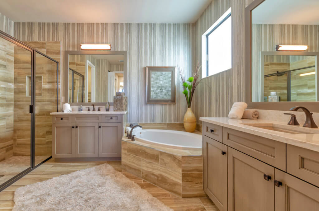 A modern bathroom at Villamar at Toscana Isles features a large glass-enclosed shower, a bathtub, and dual sinks. The striped tile walls complement a fluffy white rug on the floor. Potted plants and neatly rolled towels add decorative touches to this serene oasis.