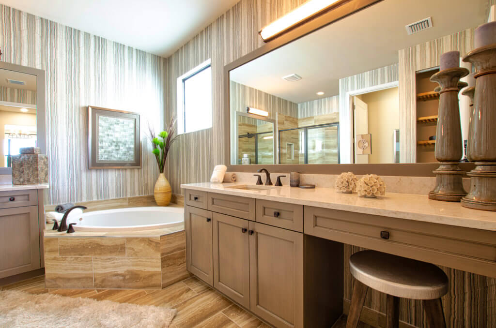 A spacious bathroom at Villamar at Toscana Isles features a large mirror above a double-sink vanity. A corner bathtub with a decorative vase adds elegance. The room boasts a beige and brown color scheme, striped wallpaper, and wooden cabinets, creating a serene oasis.