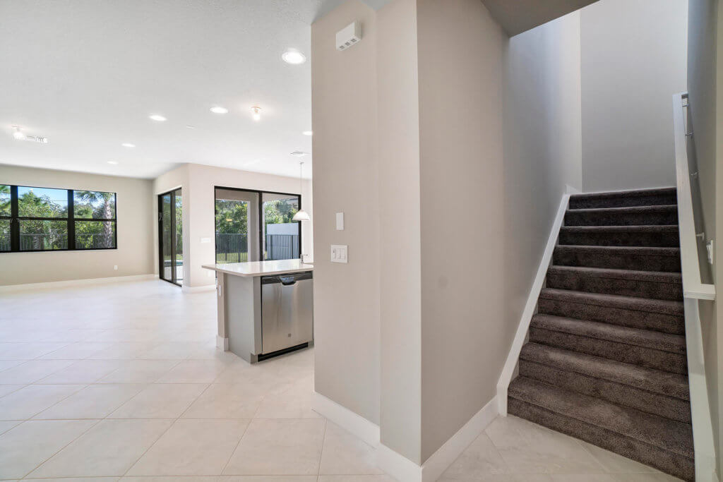 A modern home interior at Villamar at Toscana Isles with an open floor plan features a kitchen island and stainless steel dishwasher. Large windows provide natural light, while carpeted stairs lead to the upper floor. Light-colored tile flooring is throughout the space.