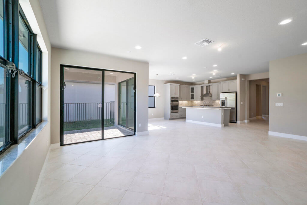 Experience the spacious charm of Villamar at Toscana Isles. This empty room boasts large windows that flood the space with natural light, complemented by light-colored tile floors and beige walls. A sliding glass door opens to an outdoor area, seamlessly connecting to a modern kitchen with stainless steel appliances.