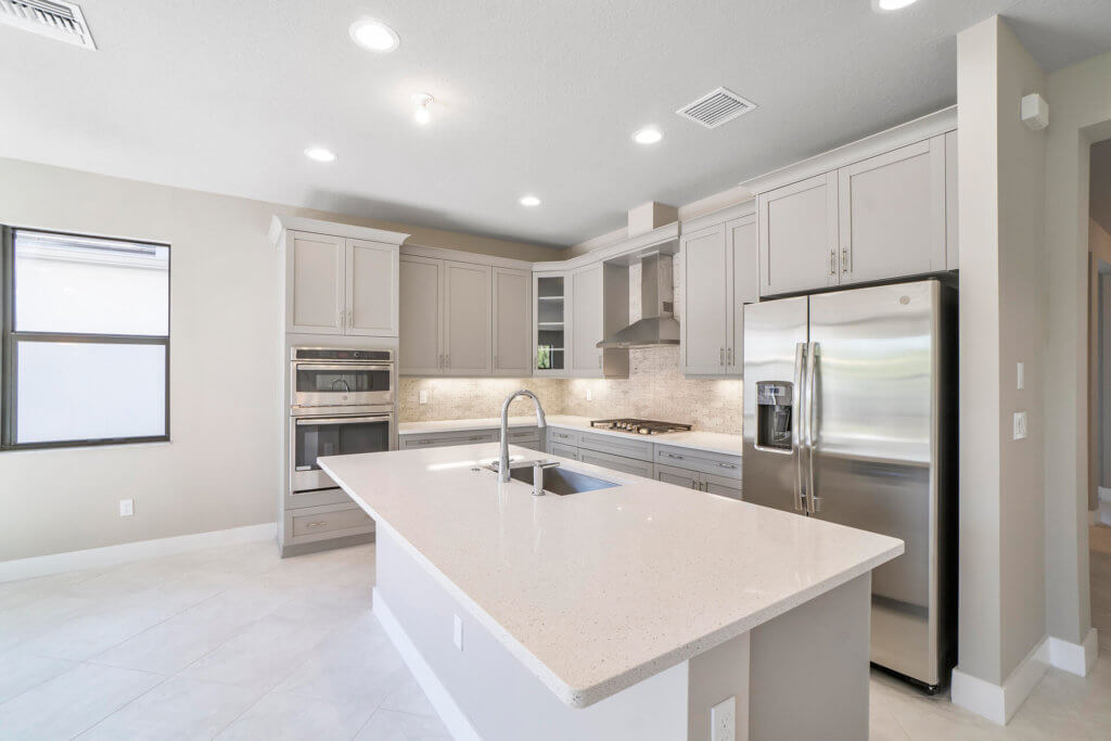 A modern kitchen at Villamar at Toscana Isles boasts white cabinets, a large center island with a sink, and stainless steel appliances, including a refrigerator, double oven, and range hood. Recessed lighting brightens the space, complemented by a window on the left.