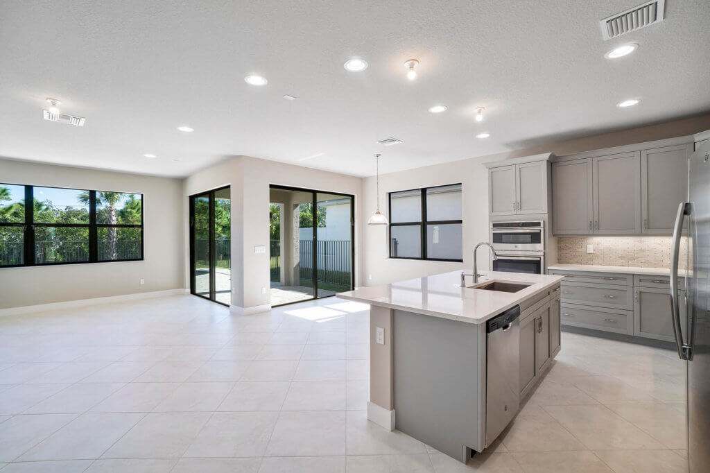 Experience the contemporary elegance of Villamar at Toscana Isles in this modern kitchen and living area, featuring light tile flooring, sleek gray cabinets, a stainless steel refrigerator, built-in ovens, and a central island with a sink. Expansive windows and sliding doors open to lush outdoor greenery.