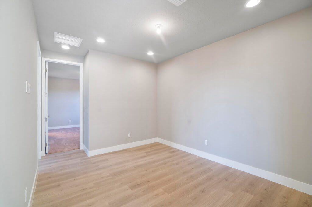 The empty room at Villamar at Toscana Isles boasts light wood flooring and beige walls. Recessed lighting and a central fixture adorn the ceiling, while a door on the left reveals another carpeted space. The area remains unfurnished, offering a blank canvas for personalized touches.