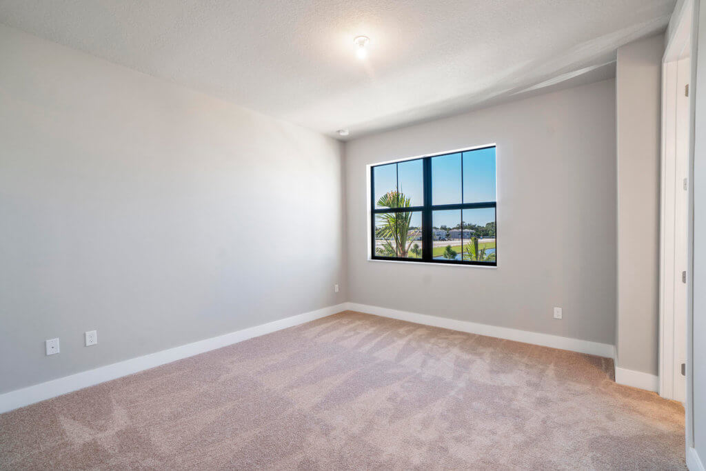 The room at Villamar at Toscana Isles boasts a bright, open space with beige carpet and light gray walls. A single ceiling light fixture illuminates the area. A large window offers a picturesque view of greenery and the serene body of water outside, highlighting the clean and well-lit ambiance.