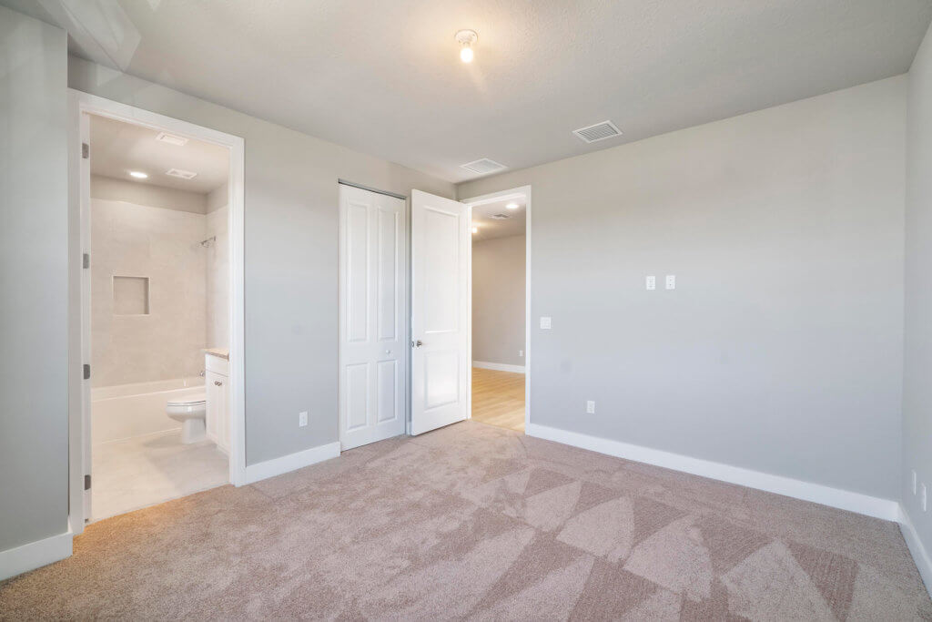 A carpeted room with neutral-colored walls in Villamar at Toscana Isles showcases an open door leading to a walk-in closet and another to a bathroom. The bathroom features tile flooring and a visible toilet, all under a ceiling adorned with an elegant light fixture.