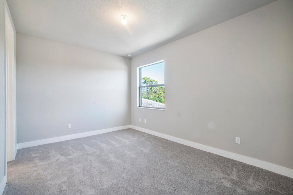An empty room with light gray walls and a single window, reminiscent of the simplicity found at Villamar at Toscana Isles. The floor is carpeted in a similar gray shade. A ceiling light is on, and there are electrical outlets on the walls. Sunlight streams peacefully through the window.