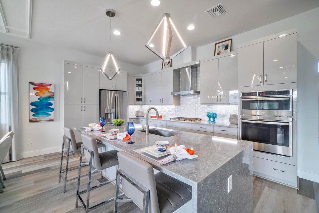 Experience the sophistication of a modern kitchen at Villamar at Toscana Isles. A large island with a gray countertop is complemented by four metallic bar stools. White cabinetry, stainless steel appliances, pendant lighting, and wall art enhance the sleek and contemporary ambiance.