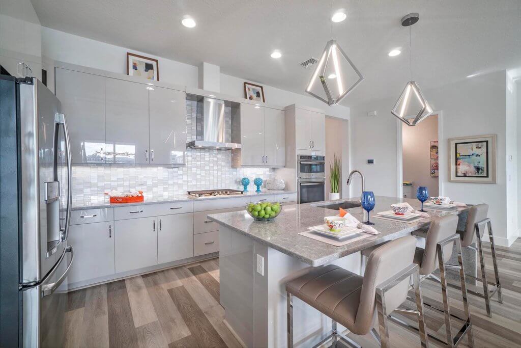 The modern kitchen at Villamar at Toscana Isles features glossy white cabinets, stainless steel appliances, and a central island with four bar stools. Geometric pendant lights hang above, illuminating place settings and glasses, while a backsplash of white and gray tiles adds elegance.