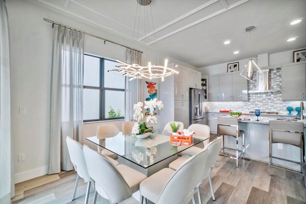 A modern dining area in Villamar at Toscana Isles boasts a glass table with beige chairs. An ornamental light fixture, adorned with white flowers, hangs above. The open kitchen features stainless steel appliances, barstools, and pendant lights, with light gray cabinets lining the walls.