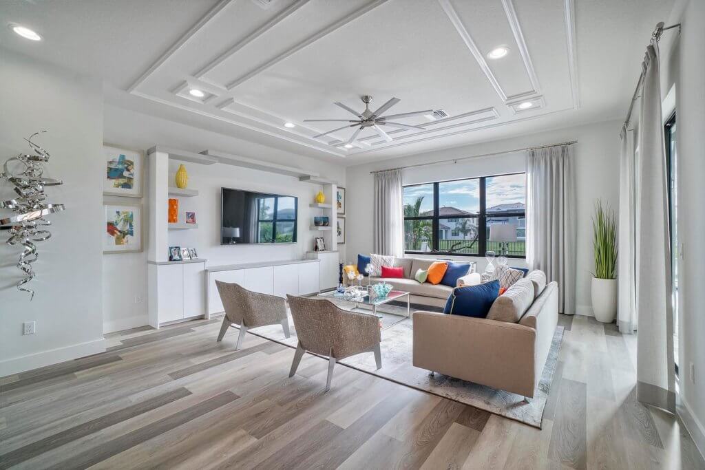 A modern living room at Villamar at Toscana Isles offers a large sectional sofa and two grey chairs facing a wall-mounted TV. The space features a coffered ceiling, wide window views, contemporary decor, and art pieces on the walls. Light wood flooring completes this elegant setting.