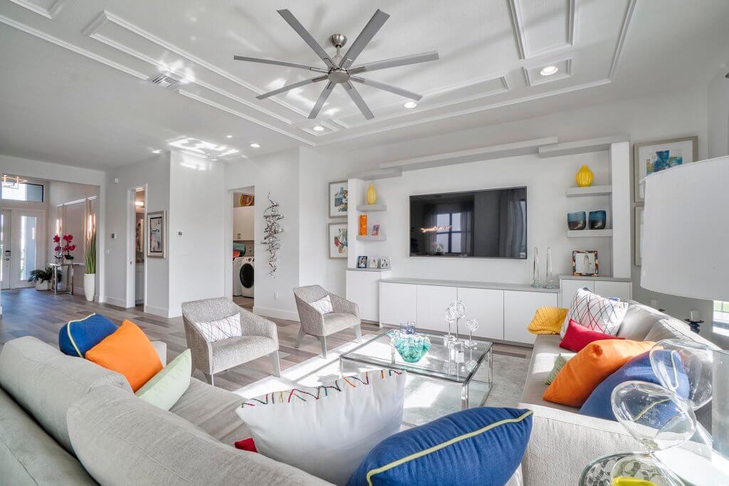 The Villamar at Toscana Isles living room boasts a modern design with a large ceiling fan, wall-mounted TV, and white shelves showcasing decorative items. A gray sofa with colorful cushions, two gray armchairs, and a glass coffee table complete the inviting space.