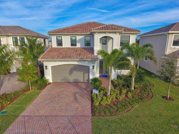 A two-story Villamar at Toscana Isles house boasts a red-tiled roof and white exterior, complemented by a brick driveway and a two-car garage. The landscaped front yard, adorned with palm trees and shrubs, completes the picturesque setting under the clear blue sky.