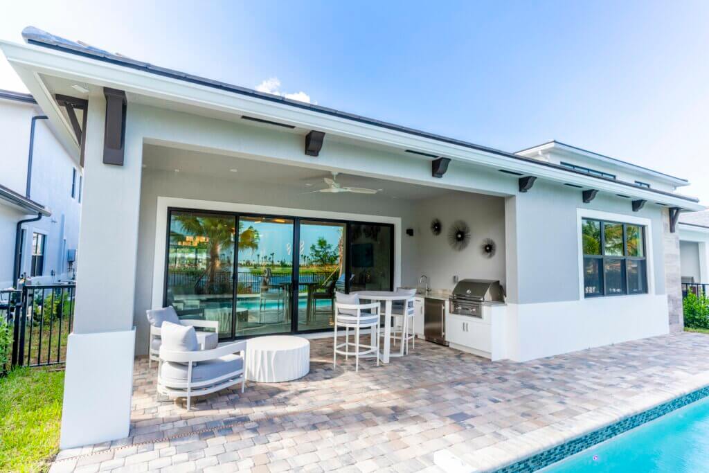 A modern patio at Solana Bay at Avenir features a lounge chair, round table, and bar-style seating. It boasts a built-in grill and sliding glass doors opening to the interior. The area overlooks a fenced yard and pool, with the sky clear and blue.