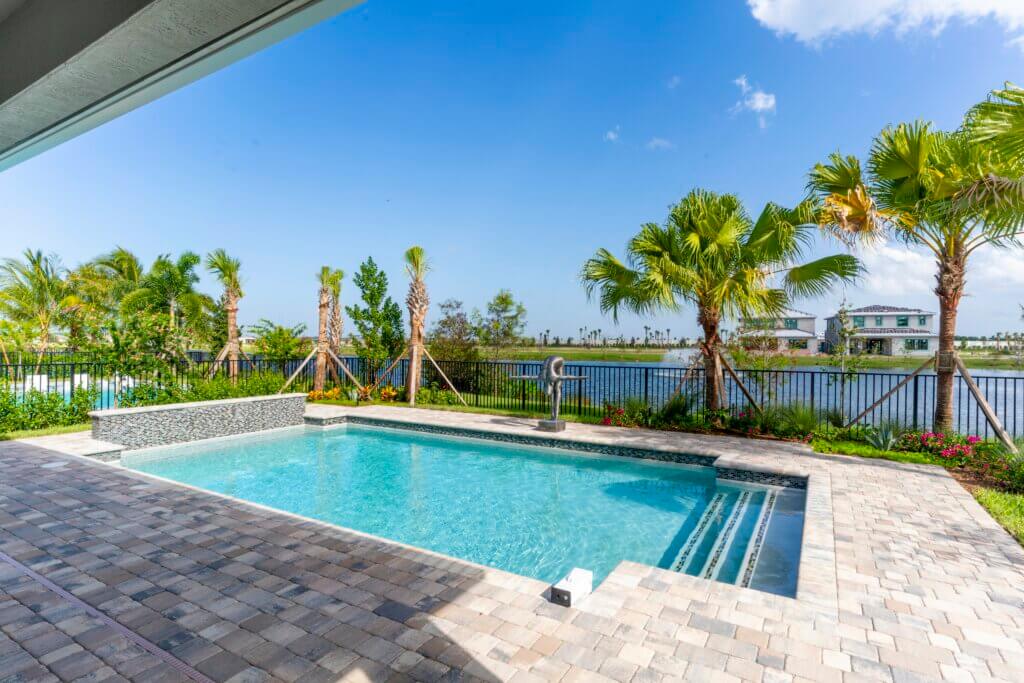 Nestled in a backyard, a rectangular swimming pool with clear blue water glistens under the sun. Surrounded by a stone patio, lush palm trees, and vibrant plants, it offers tranquility. A fence outlines the area, unveiling views of Solana Bay at Avenir in the distance.