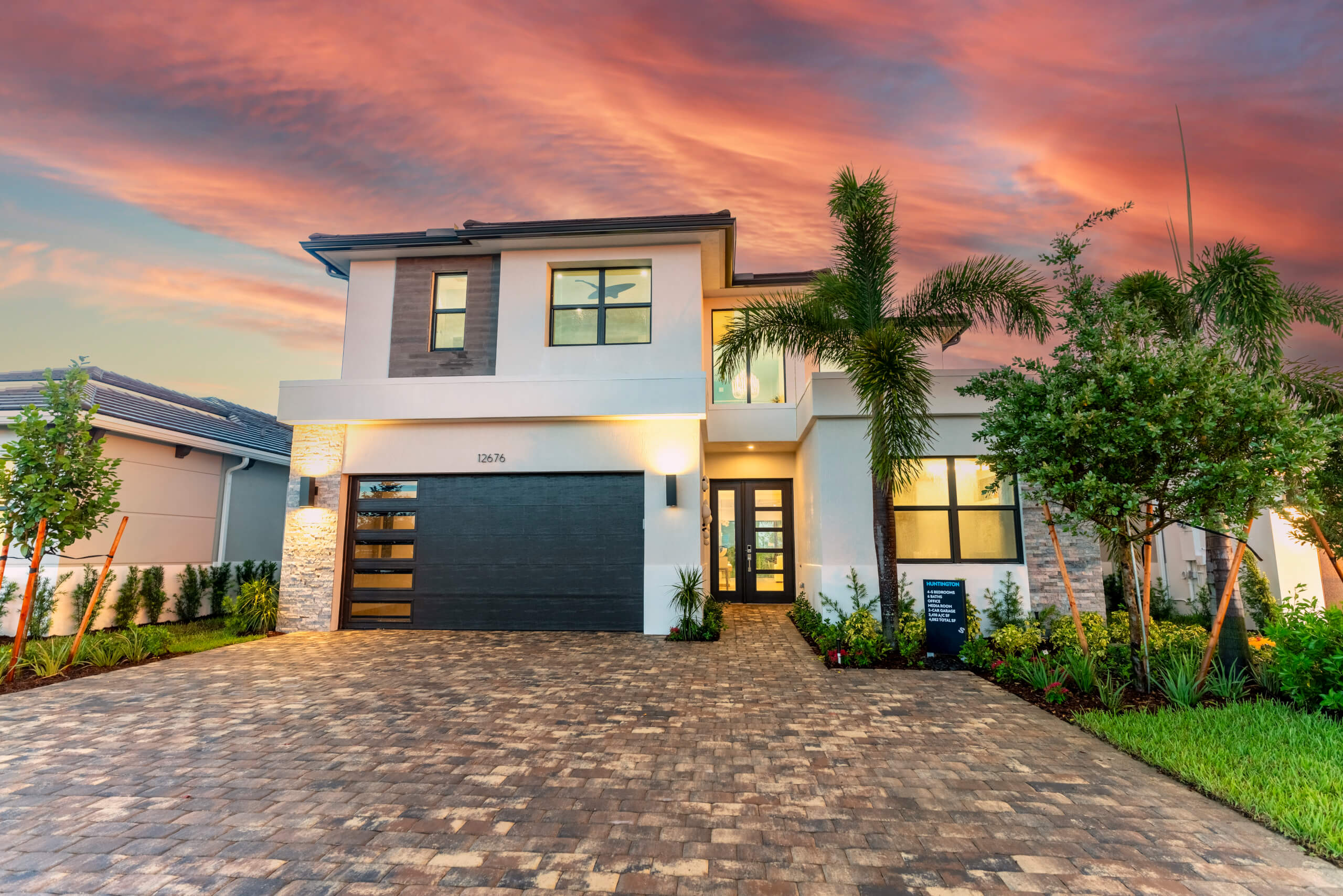 Nestled in Solana Bay at Avenir, this modern two-story house boasts a brick driveway and garage, set against a dramatic sunset sky. With large windows, palm trees, and lush greenery adorning the property, it offers a serene retreat amidst nature's splendor.