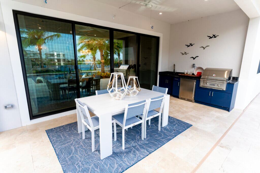Outdoor patio at Solana Bay at Avenir features a modern white dining table with four chairs on a blue patterned rug. Large glass doors reflect palm trees and a pool, while the adjacent kitchen area boasts blue cabinets, a grill, and bird decorations on the wall.