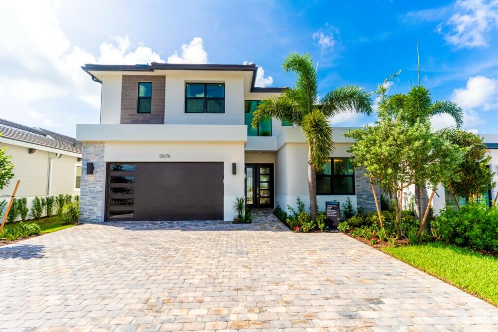 A modern two-story house in Solana Bay at Avenir boasts a chic mix of white and gray exterior walls, large windows, and a dark garage door. The driveway features light-colored bricks, with palm trees and manicured shrubs adorning the front yard under a clear blue sky.