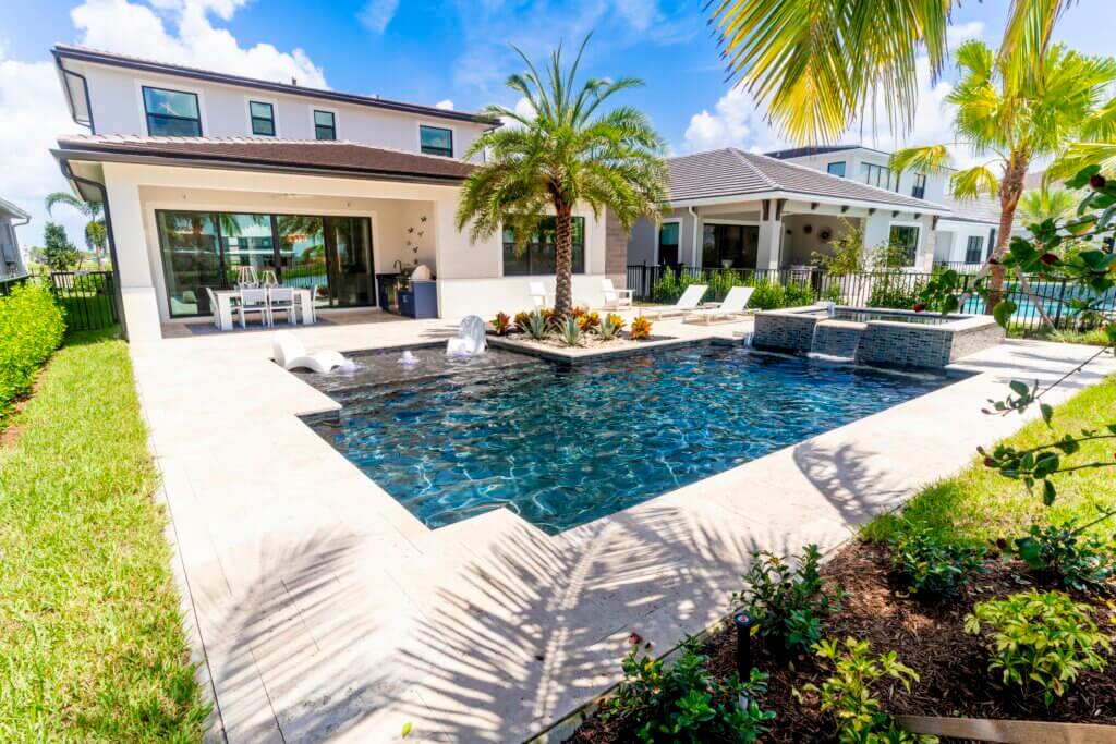 A modern backyard at Solana Bay at Avenir features a rectangular swimming pool surrounded by a stone deck. Lounge chairs, a dining table, and tropical plants, including a tall palm tree, enhance the scene. In the background is a two-story house with large windows and a covered patio.
