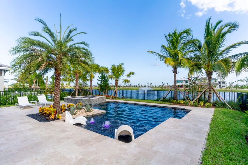 This luxury backyard at Solana Bay at Avenir boasts a rectangular pool encircled by a stone patio. Palm trees line the area, where two white lounge chairs rest poolside. A black fence frames the serene lake view with distant structures beyond.