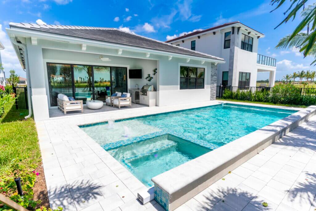 A modern house with large windows overlooks the clear blue pool of Solana Bay at Avenir, surrounded by a white-tiled patio adorned with outdoor furniture. In the background, a second-story building stands under the bright blue sky dotted with a few clouds.