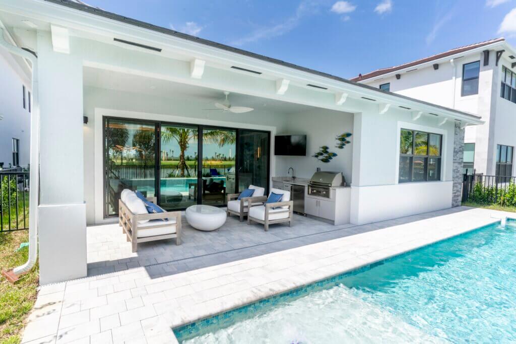 The modern outdoor patio at Solana Bay at Avenir showcases a seating area with gray furniture and blue cushions, adjacent to the inviting swimming pool. A built-in grill and wall-mounted TV are sheltered under a covered roof, while large glass doors reflect the palm trees beautifully.