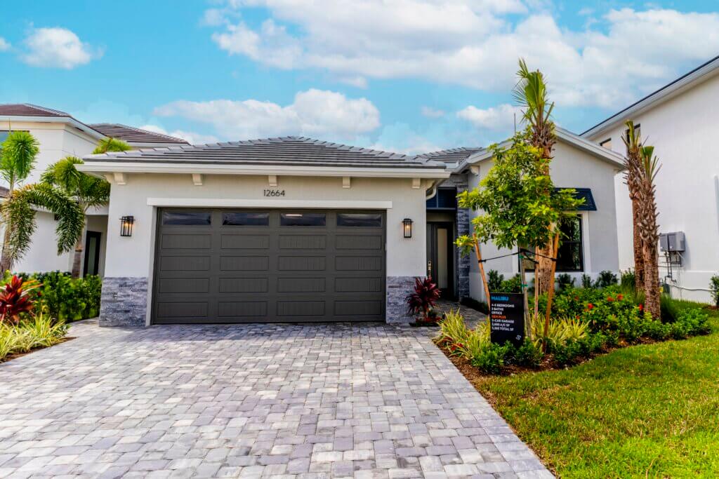 A modern single-story house in the sought-after Solana Bay at Avenir, featuring a gray tiled roof, two-car garage, and pristine white walls. The front yard boasts a stone driveway, landscaped plants, and lush lawn. A real estate sign stands nearby under the partly cloudy sky.