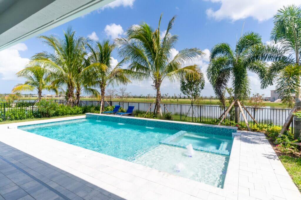 The backyard scene at Solana Bay at Avenir features a rectangular swimming pool with clear blue water. Palm trees and lush greenery line the poolside, offering a serene escape. Beyond the pool, a tranquil view of the lake complements the clear sky dotted with a few clouds.