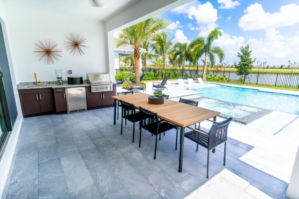 The outdoor patio at Solana Bay at Avenir features a long wooden dining table with black chairs under a covered area. To the side, there's a built-in grill and countertop. Beyond, a pool with lounge chairs and palm trees overlooks a fenced grassy area under a blue sky with clouds.