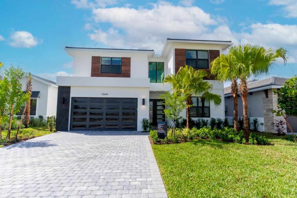 A modern two-story house in Solana Bay at Avenir boasts a white exterior, dark garage door, and elegant wooden accents. The front yard features a paved driveway, lush green lawn, and graceful palm trees under a partly cloudy sky.