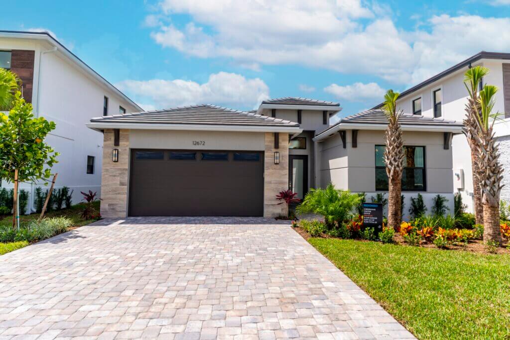 A modern single-story house at Solana Bay at Avenir features a gray tiled roof, stone accents, and a double garage door. The light-colored brick driveway and pathway complement the neatly landscaped plants and green lawn surrounding this exquisite home.