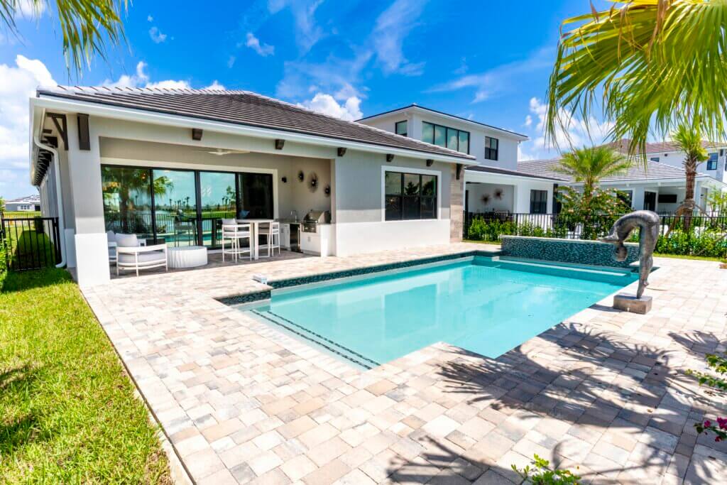 The modern backyard at Solana Bay at Avenir features a swimming pool surrounded by stone tiles. A patio with a dining set nestles under a covered area, attached to a house with large glass doors. In the background, another building and palm trees are visible under the blue sky.