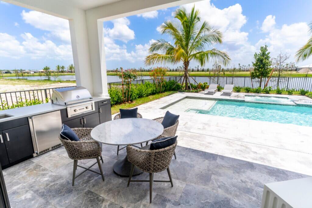 The outdoor patio at Solana Bay at Avenir features a round marble table and four wicker chairs, complemented by a built-in stainless steel grill. This serene area overlooks a swimming pool, palm trees, and the scenic view of a lake beneath a partly cloudy sky.