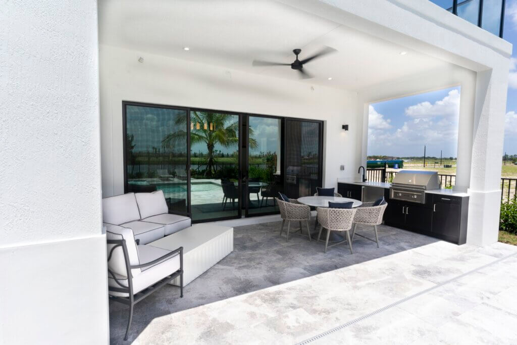 The modern outdoor patio at Solana Bay at Avenir boasts a white cushioned sofa, a sleek coffee table, and a round dining table with chairs. A ceiling fan spins above, while a built-in grill awaits on the right. Large sliding glass doors reflect the tranquil pool and lush palm trees beyond.