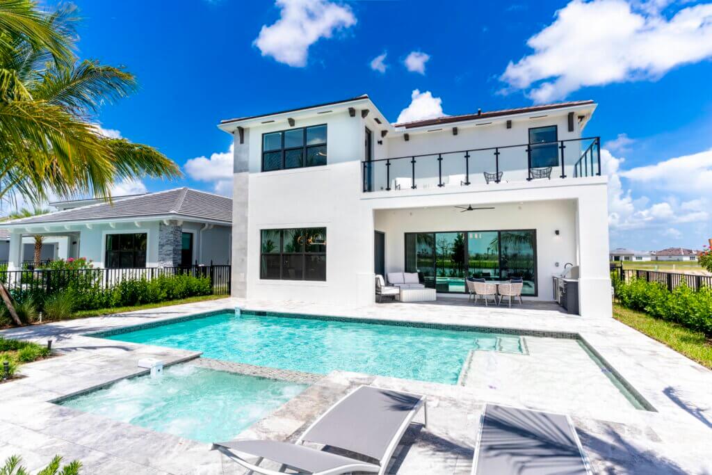 This modern two-story house in Solana Bay at Avenir features large windows, a balcony, and a covered patio. In the foreground, there's a swimming pool with a hot tub surrounded by a stone deck and lounge chairs. A palm tree stands to the left under a bright sky dotted with clouds.