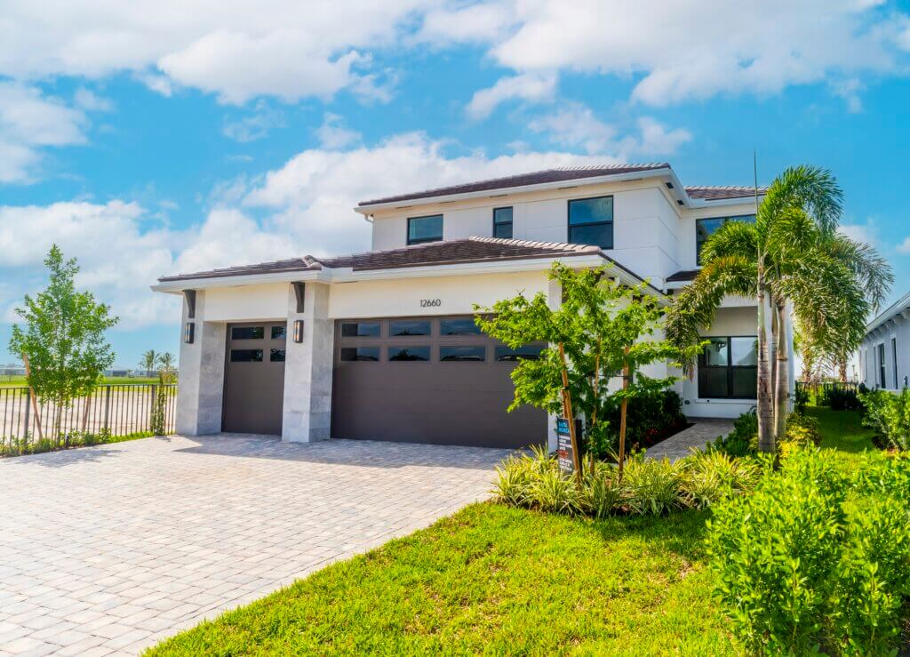 A two-story modern house in Solana Bay at Avenir boasts a tiled roof and large garage doors. Its exterior features white walls with stone accents, and a paved driveway winds through landscaped greenery and palm trees. The sky above is partly cloudy, adding to the serene ambiance.