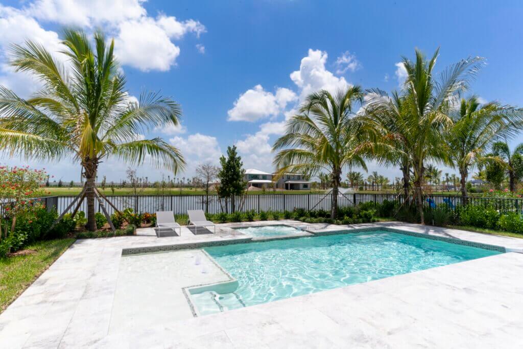 The backyard at Solana Bay at Avenir showcases a rectangular swimming pool with a small attached hot tub, set amidst a stone patio. Two lounge chairs rest poolside, while palm trees border the fence. In the distance, a shimmering body of water is visible under the expansive blue sky.