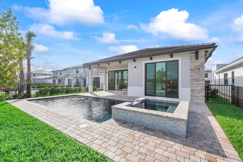 Modern house with large windows, a tiled patio, a rectangular swimming pool, and a hot tub, surrounded by green grass and a black metal fence under a blue sky.