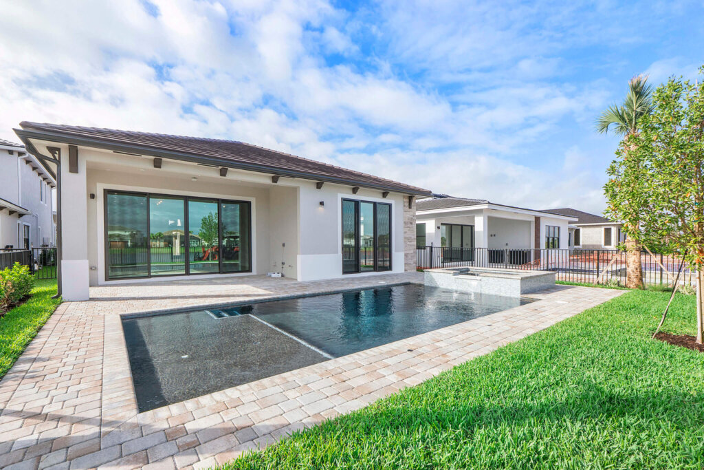 Modern single-story house with large sliding glass doors, a rectangular swimming pool, paved patio, and green lawn under a partly cloudy sky.