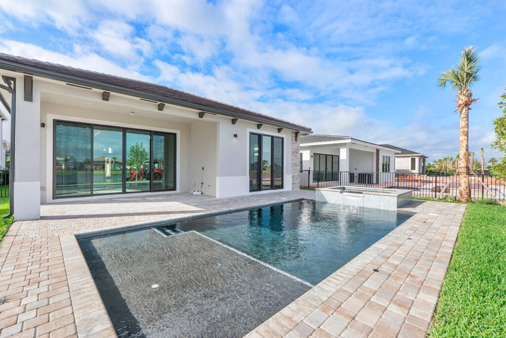 Modern house with large glass doors and a rectangular backyard pool surrounded by stone pavers, next to a grass lawn and palm tree, under a partly cloudy sky.