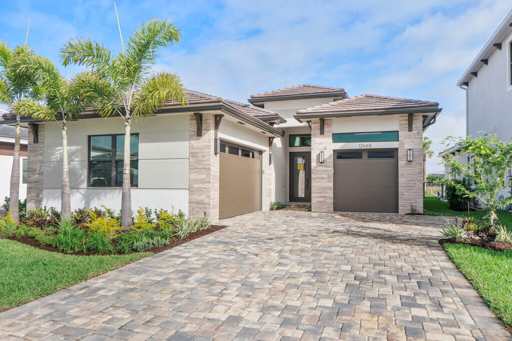 Single-story modern house with two garages, a paved driveway, and landscaped front yard with palm trees under a partly cloudy sky.