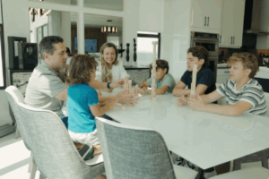 A family of six sits around a white dining table playing a stacking block game in a modern kitchen.