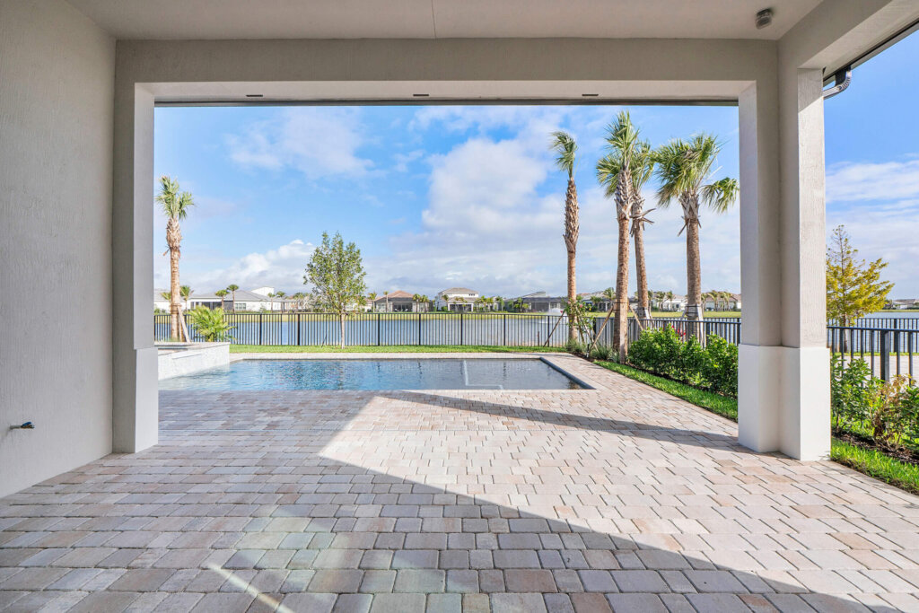 Covered patio with paver flooring overlooking a pool, landscaped yard, and a lake with houses and palm trees in the background under a partly cloudy sky.