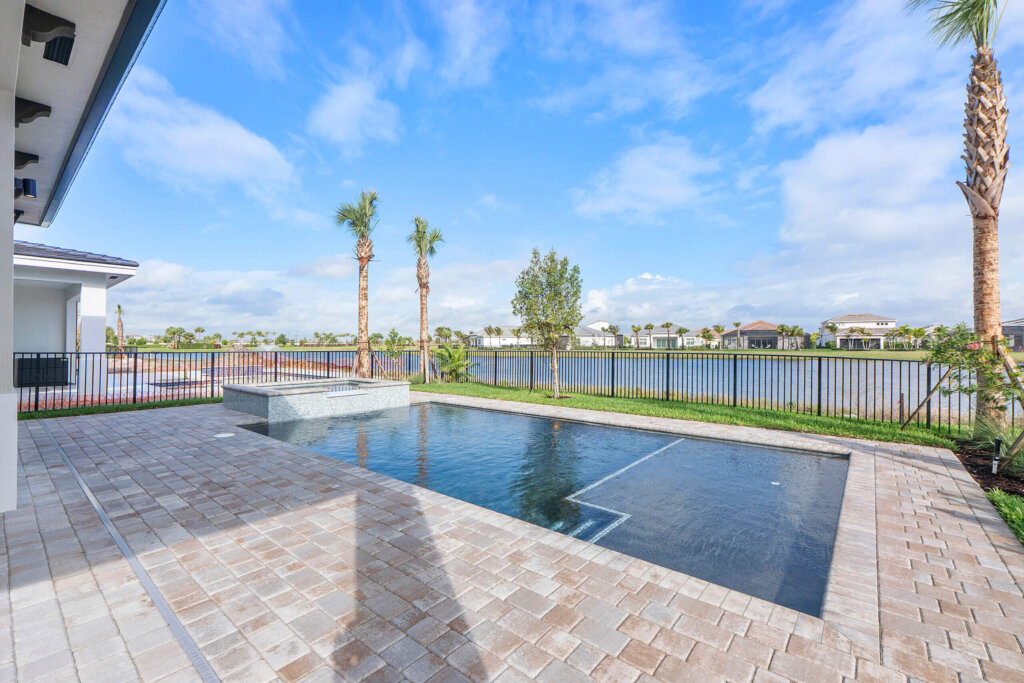 Rectangular backyard pool with a paved patio, fenced perimeter, palm trees, and a view of a lake with houses in the background under a blue sky.
