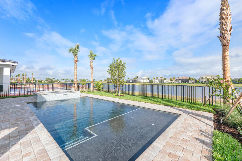 Rectangular backyard pool with a raised spa, surrounded by pavers, overlooking a lake with palm trees and houses in the background under a blue sky.