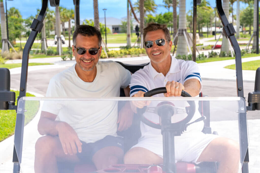 Two men sit together in a golf cart. The man on the left wears a grey shirt and sunglasses, while the man on the right, in a white polo and sunglasses, is seated at the wheel. Palm trees and a grassy area are visible in the background.