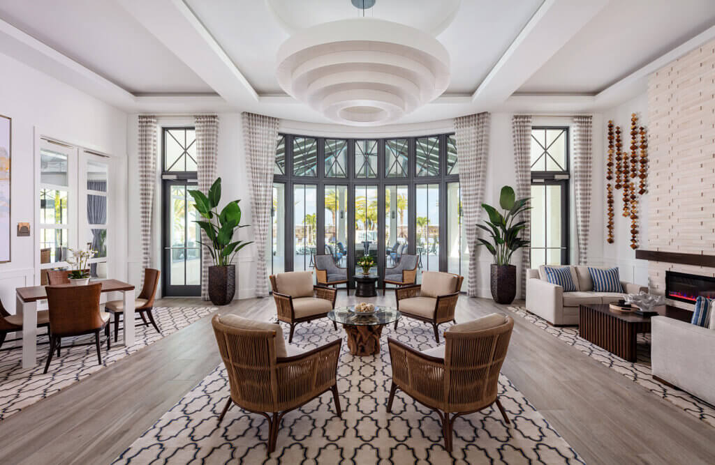 A spacious, modern living room at Catalina Palms at Sundance embraces a light color palette. It boasts a circular seating arrangement with wicker chairs around a central glass coffee table. Large windows, adorned with curtains, allow sunlight to highlight potted plants and decorative accents.