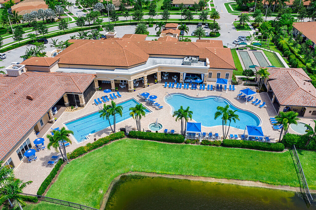 Aerial view of Catalina Palms at Sundance resort, showcasing a large, curvy swimming pool and a rectangular pool encircled by lounge chairs and blue umbrellas. The terracotta-roofed buildings are complemented by palm trees and a well-maintained lawn near the water's edge.