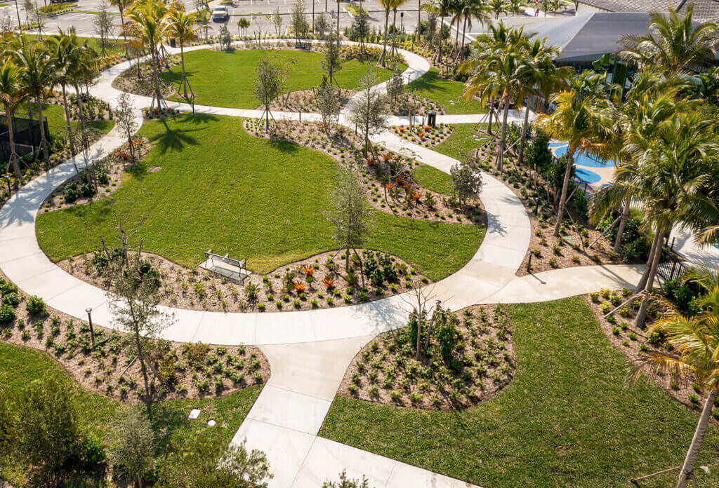 Aerial view of a landscaped park, Catalina Palms at Sundance, featuring winding pathways, green lawns, and various planted areas. Palms line the paths, and a bench sits along one walkway. The park is surrounded by a parking area and other structures.